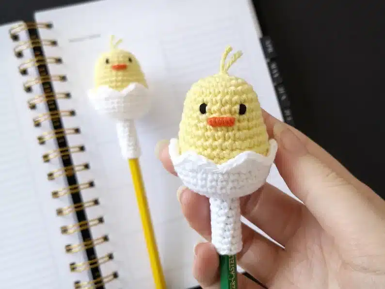 Crochet yellow chick sticking out of a white egg place on top of a green pencil, another one placed on a yellow pencil in the background. 