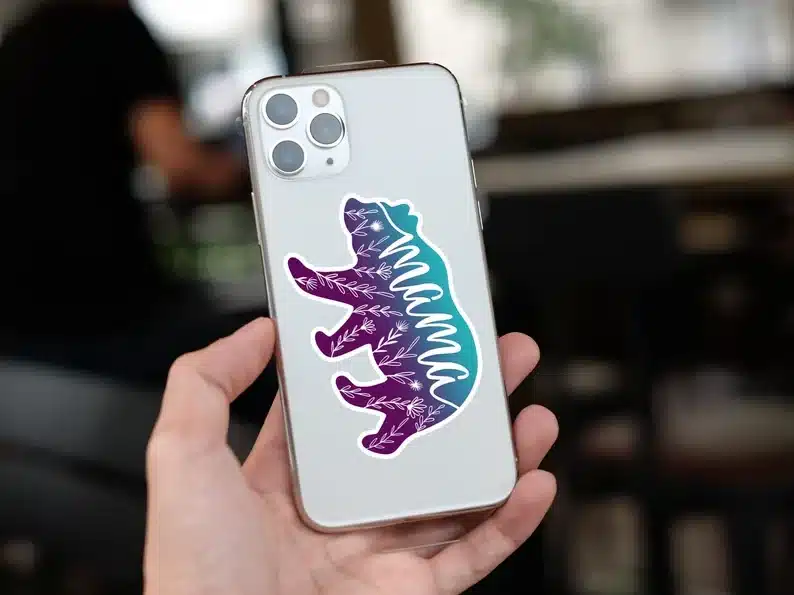 showing white phone case being held with a blue and purple bear shaped decal that says MAMA on it. 