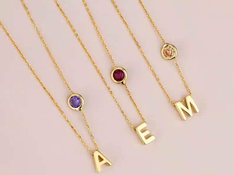 Three gold necklaces, one with an A charm and a purple birthstone gem shown, one with an E and a burgundy Gemstone shown, and lastly one with an M charm with a cream colored gemstone. 