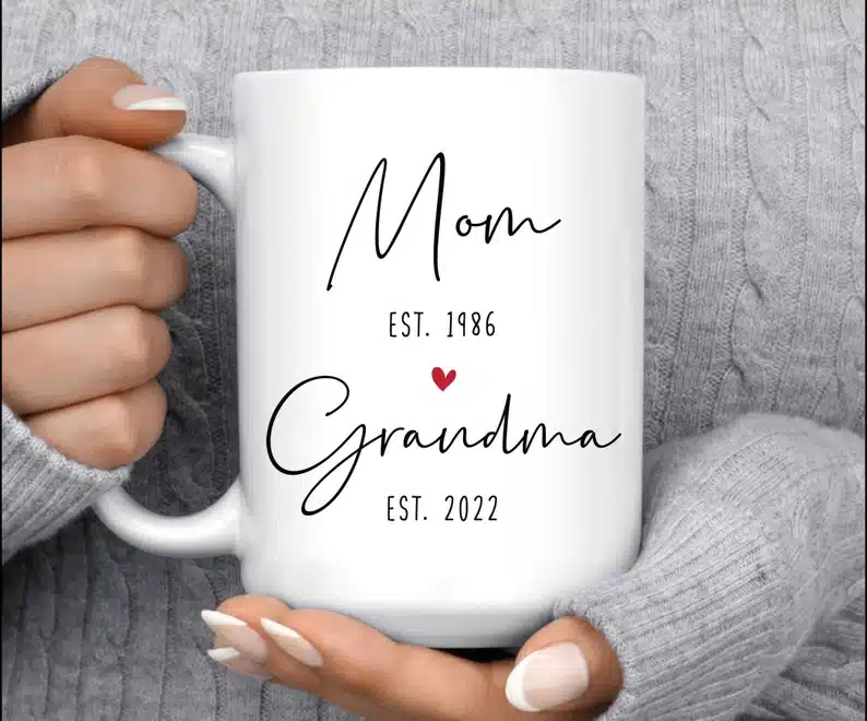 Close up of Woman's hands holding a white coffee mug with black font that says MOM est 1986. Grandma EST 2022. 