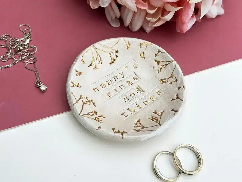 Round clay cream colored dish with gold font that says Nanny's rings and things. 