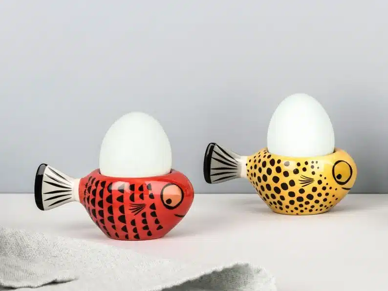 Two matching egg cups, both fish; one red with black scales and another yellow with black scales. 