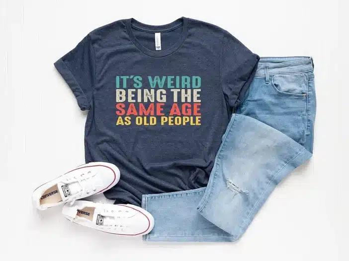 Navy blue t shirt that colorful writting that says It's weird being the same age as old people. With a pair of jeans and white sneakers beside it. 