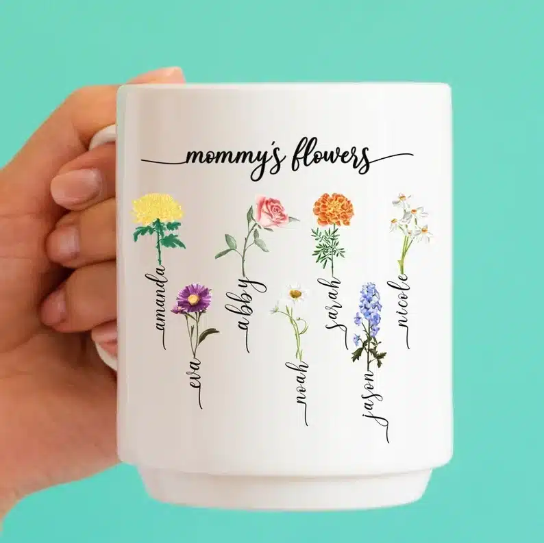 Close up of woman's hand holding a white coffee mug that says Mommy's flowers and many kids names with a flower. 