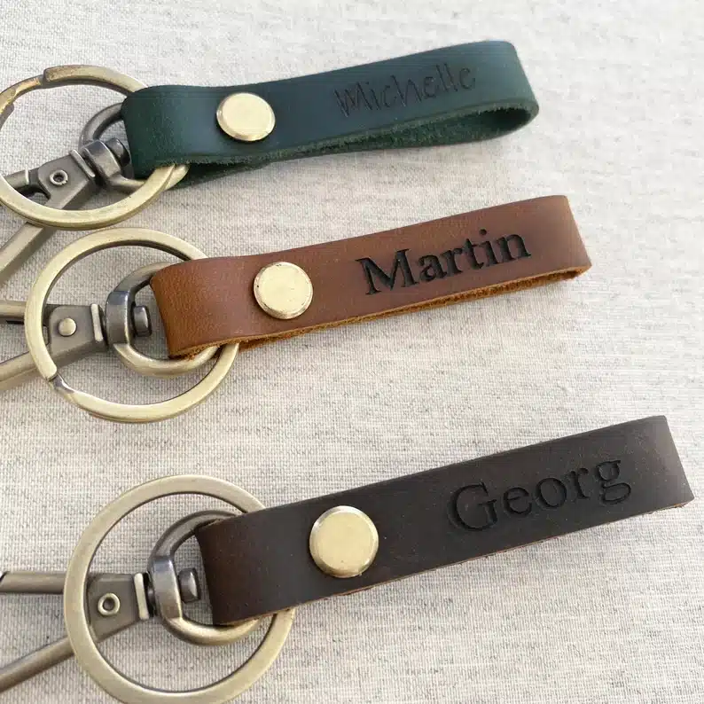 Customized leather keychains, 3 shown all different colors: teal with Michelle on it in black front, brown with Martin on it, and Georg on a grey keychain. 
