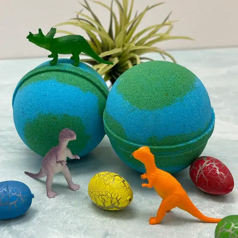 Two blue and green bath bombs with various toy dinosaurs around it and little dino eggs shown. 