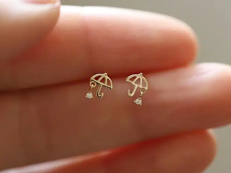 Close up of a woman fingers holding two small gold umbrella earrings with ting diamond looking raindrop. 