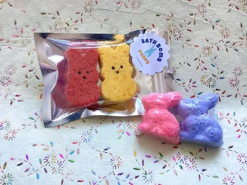 Four different Bunny shaped bath bombs, two in a clear wrapper shaped like Easter peeps one red and one yellow, and two individual wrapped full bunnies one pink and one blue. 
