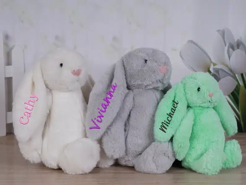 Personalized stuffed bunnies, one white with Cathy on the ear in pink, one light grey with Viviana in purple on its ear and a slightly smaller mint bunny with Michael in dark green font on the ear. 