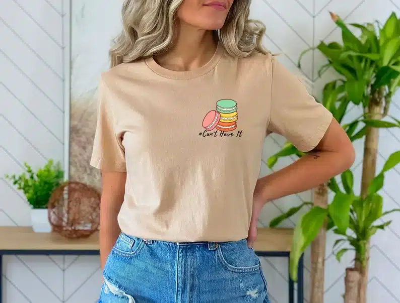 Woman wearing a cream colored t-shirt with colorful macaroons on it that says below in black font 