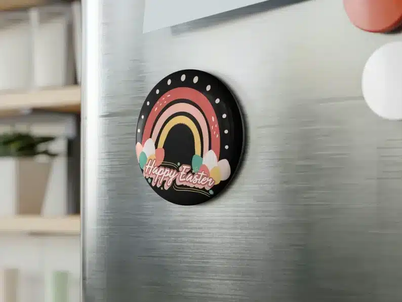 CLose up of a stainless steel fridge with a black round magnet on it that has a pink, lighter pink, and yellow rainbow and eggs on the end of each side of rainbow that says HAPPY EASTER below it. 