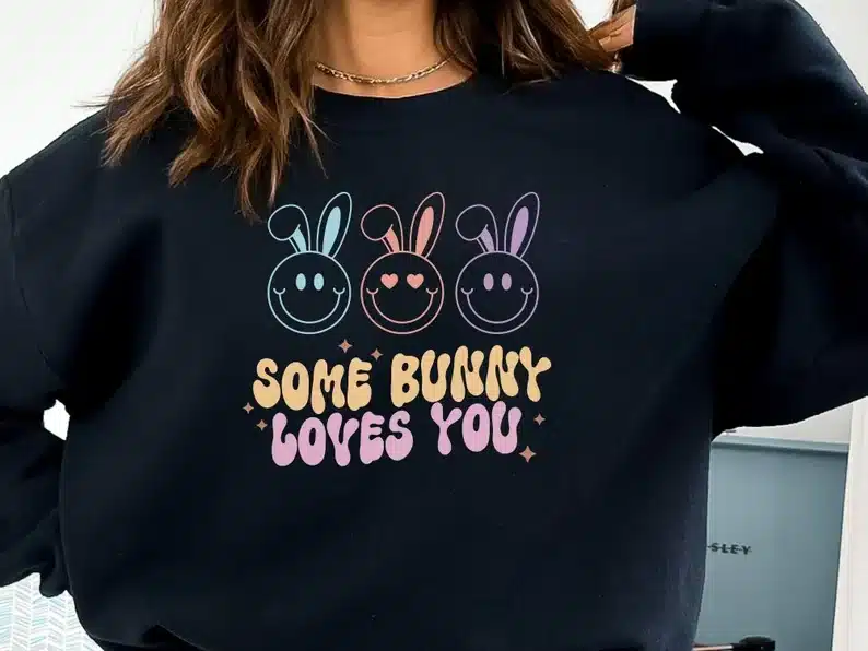 Woman wearing a black long sleeve sweatshirt with three happy faces with bunny ears and below them orange font that says SOME BUNNY and pink font below that that says LOVES YOU. 