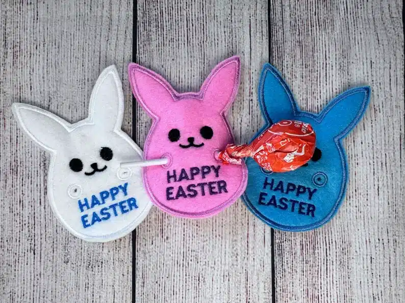 Three felt bunnies: white, pink, and blue all with Happy Easter sewn on with a orange tootsie pop sticking through them. 