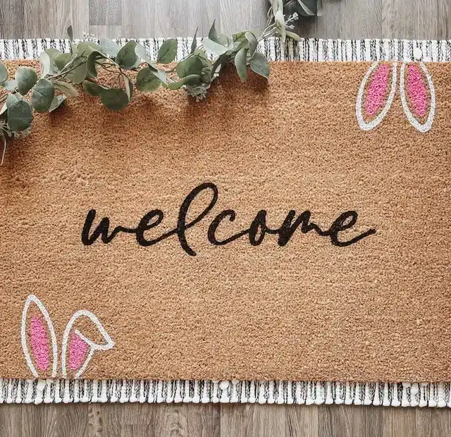 Brown mat with black font that says WELCOME with two bunny ears at the bottom and top sticking out with pink in the ears. 
