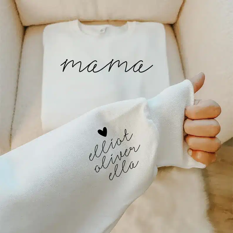 White t-shirt folded up on a cream colored couch that says MAMA with black font and a close up of a woman's arm sleeve that says Elliot, oliver, and ella on it. 