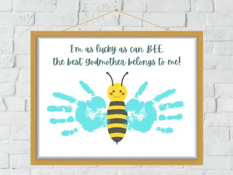 Handprint art framed in a brown frame. Showing a bee with blue handprints as wings and font above that says I'm as lucky as a bee the best godmother belongs to me! 