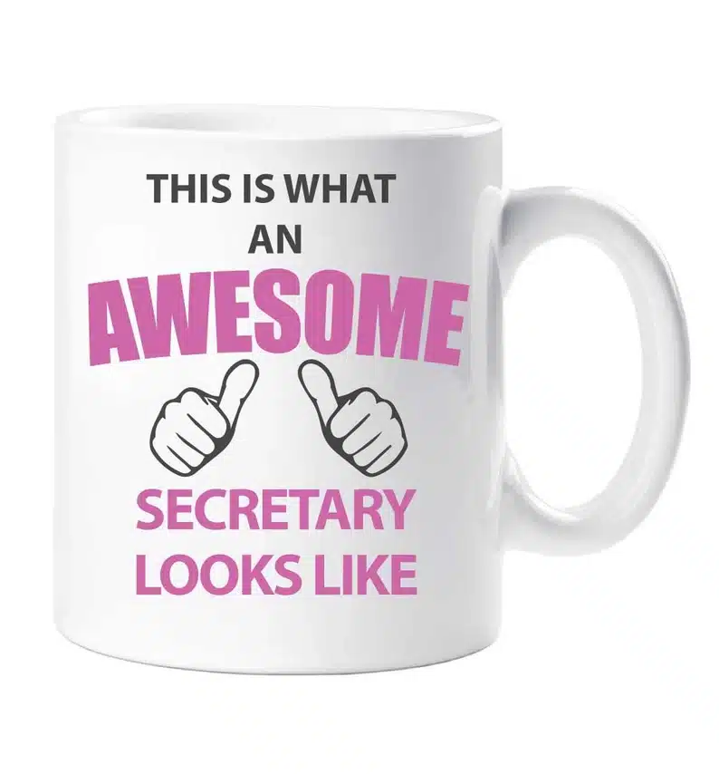 White coffee mug with black font that says "This is what an" now in pink font "awesome secretary looks like" with two thumbs pointing up. 