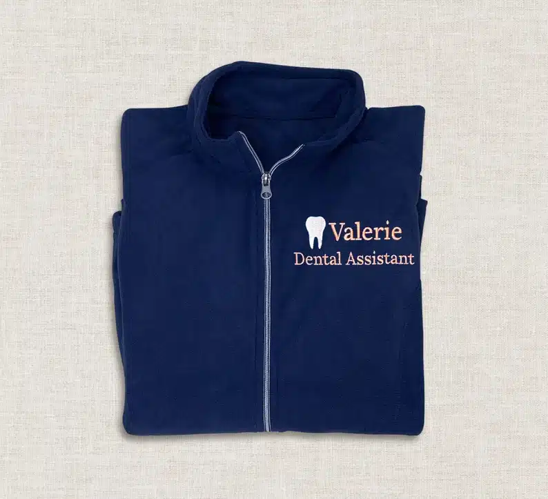 Gift Ideas For Dental Assistants: Custom embroidered dental assistant jacket. Navy blue jacket shown with a stitched on white tooth with Valerie Dental assistant in pink stitched beside tooth. 