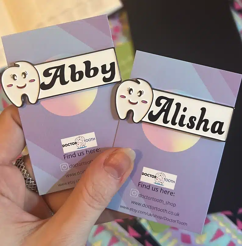 Gift Ideas For Dental Assistants: Close up of a woman hands holding two custom name tags, both with a smiling tooth and a name in black font beside it. 