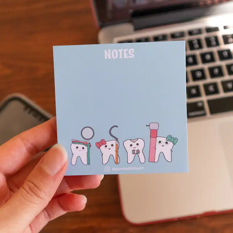 Close up of hand holding a light blue sticky notepad with four different cartoon teeth at the bottom. 
