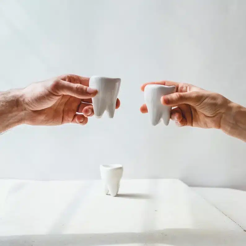 Gift Ideas For Dental Assistants: two hands each holding a tooth shaped shot glass, with one shot glass below on the table. 