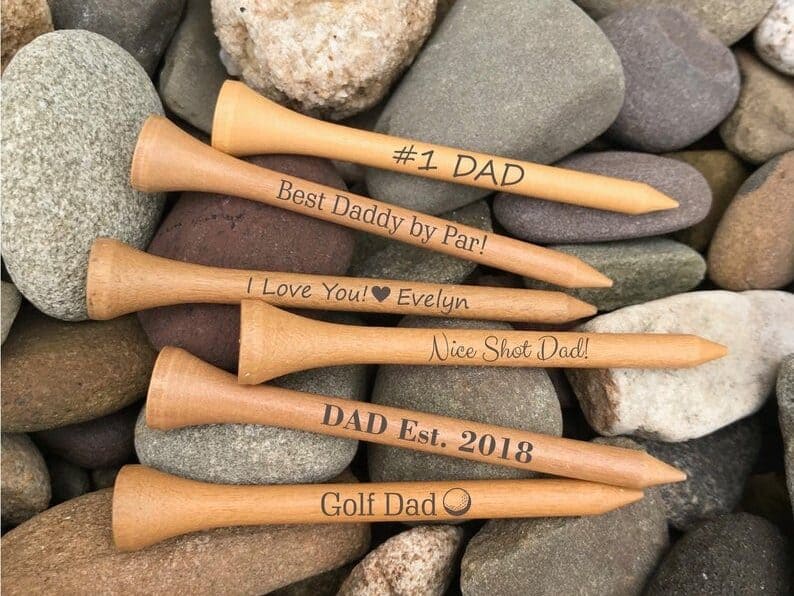 Six wooden golf tees laying on top of rocks, each with a saying about dad on them. 