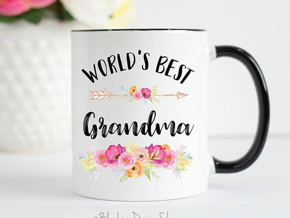 White coffee mug with a black handle/ Black font that says "world's best grandma" with flowers on it.