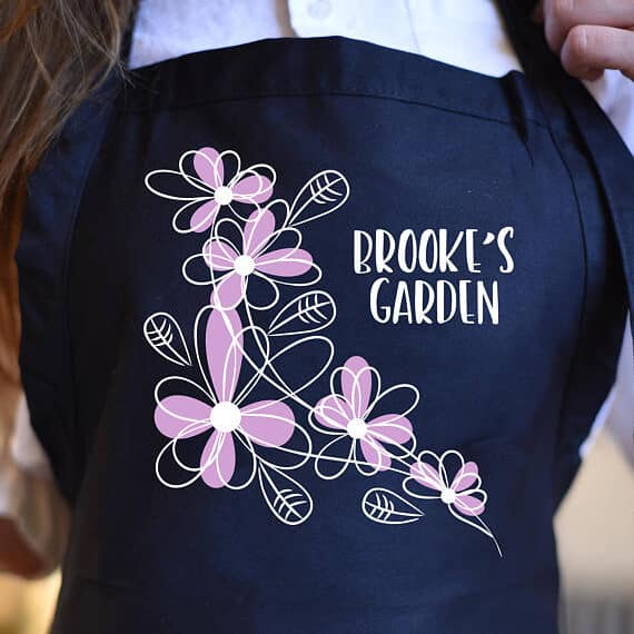 Close up of a black apron with white font that says Brooks garden and pink flowers beside it.