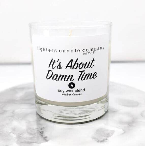 Glass jar with a white candle in it with black font that says It's about damn time.