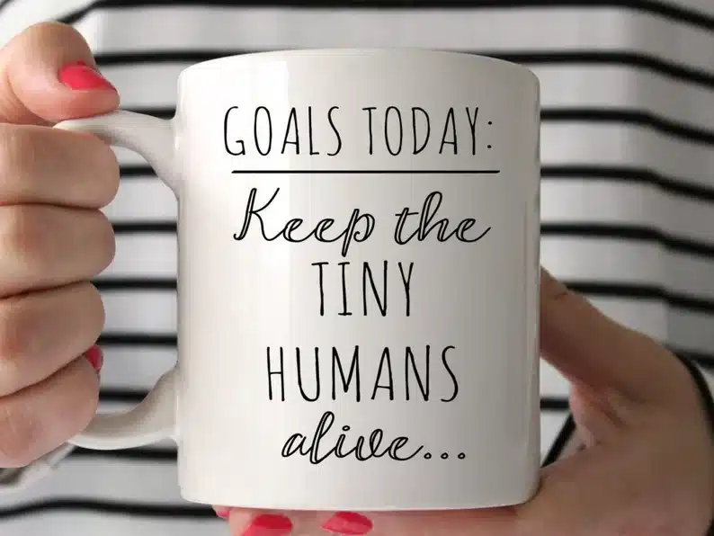 Close up of woman's hands holding a white coffee mug with black font that says "goals today: keep the tiny humans alive...
