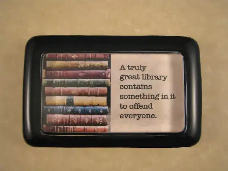 National Library Workers Day Gift Ideas: Black paperweight that shoes books and black font that says "A truly great library contains something in it to offend everyone." 
