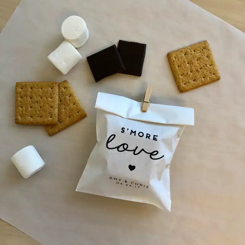 Welcome Bag Gifts for Destination Weddings: white package with black font that says s'more love. with graham crackers, chocolate and marshmallows laying around white bag. 