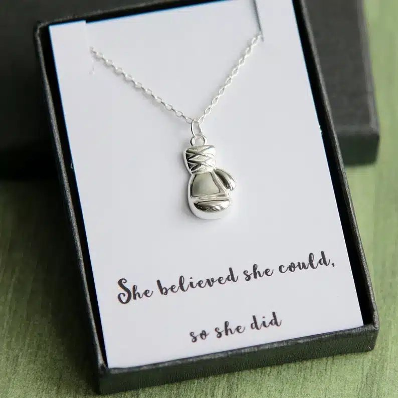 black box with a white inset with a silver boxer glove charmed necklace in it. Black font below necklace that says "She believed she could, so she did" 