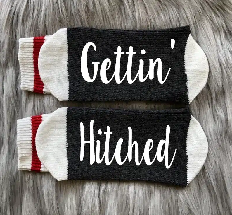 Dark grey socks with white tips and a red strip on the top. White font on each sole that says "Gettin'" on one and "Hitched" on the other. 