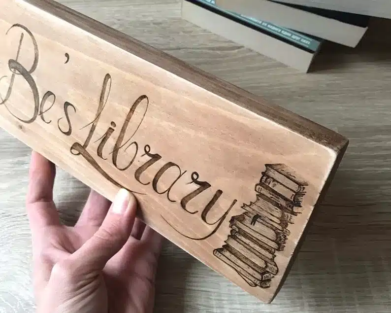 Wooden rectangle engraved customable sign that says Be's Library" with a stack of books. 