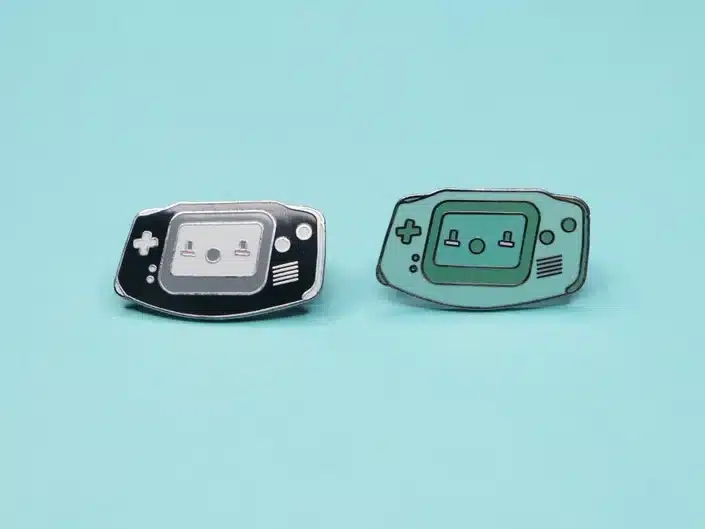 Two Gameboy enamel pins, one black and white and one teal and darker teal. 