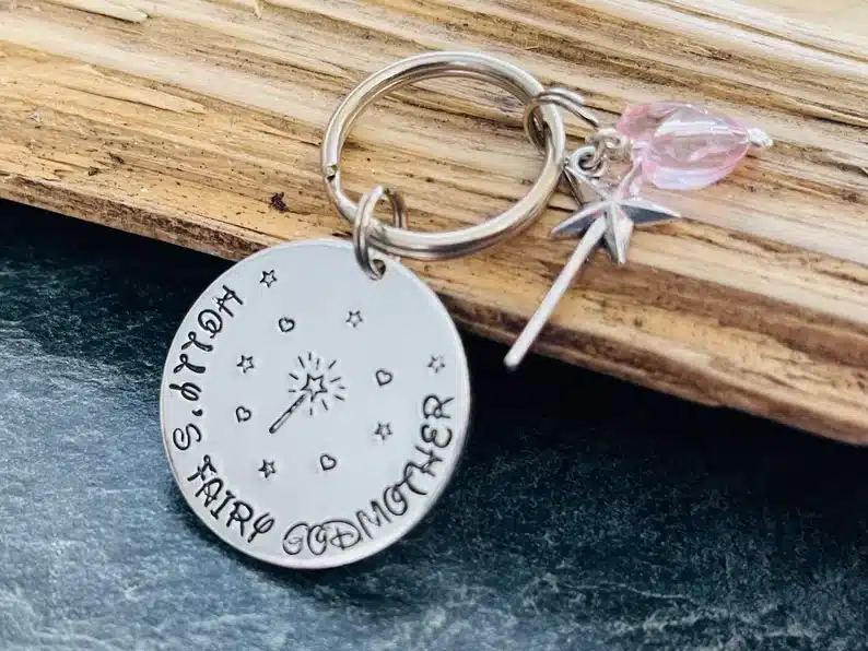 Silver keychain with a round charm that says holly's fairy godmother" and a star wand charm beside it. 