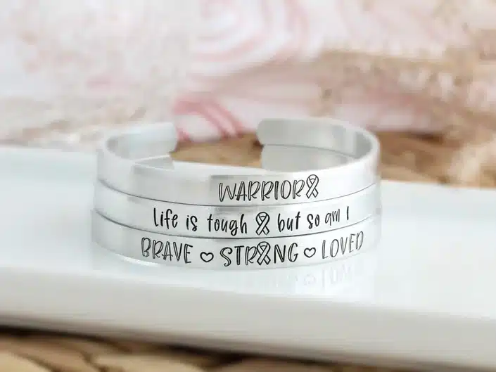 Mother's Day Gifts for Cancer Patients - Three silver cuff bracelets, all with black ink that says Warrior, life is tough but so am i and lastly brace strong and loved. 