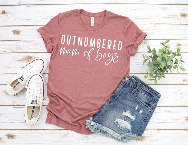 Dusty rose t-shirt with white font that says "Outnumbered mom of boys" with jean shorts and white shoes beside it.