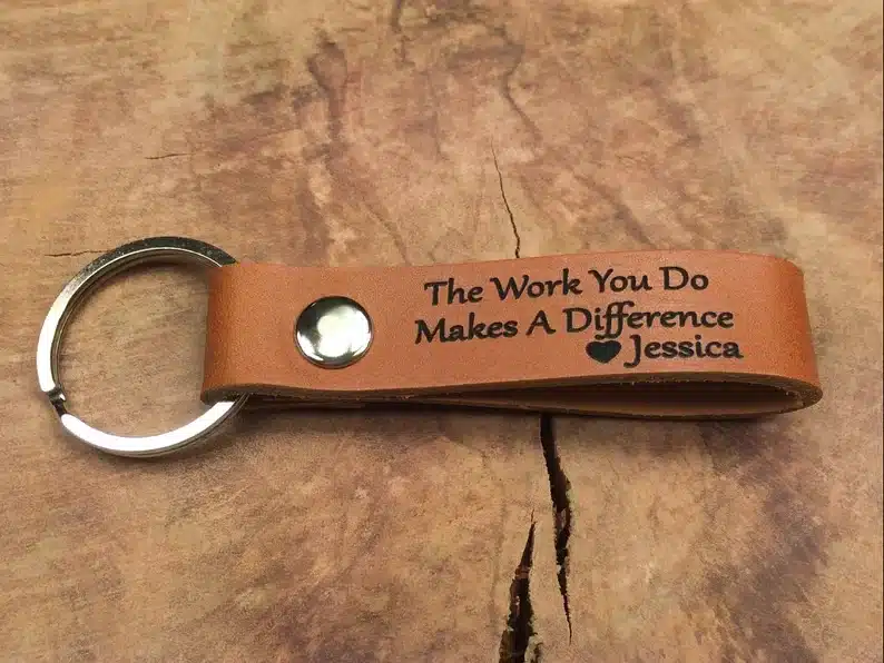 Silver key ring with light brown leather attachment with black font that says "the work you do makes a difference" 