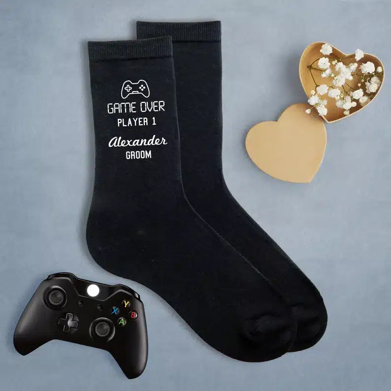 Black socks with white font that says Game over player 1 Alexander Groom with a game controller on them. 