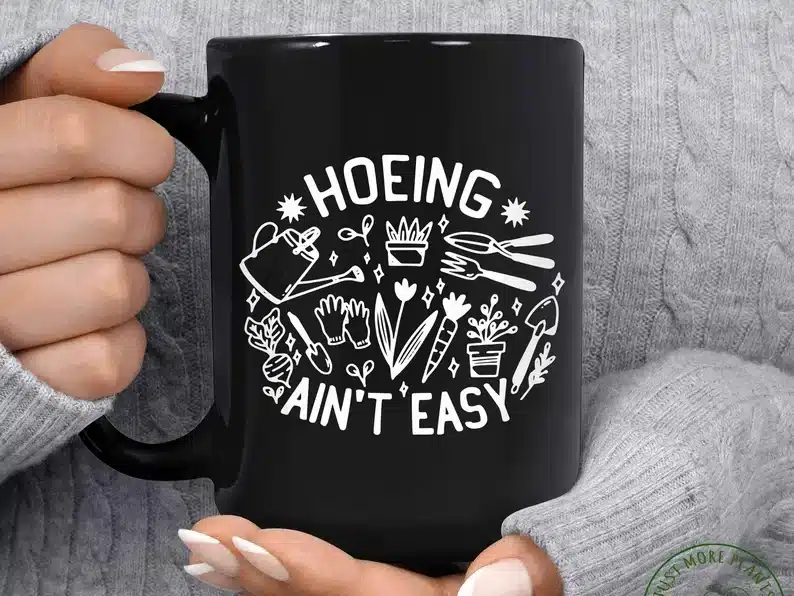 Mother’s Day Gifts for Gardeners: Black coffee mug with white font that says Hoeing ain't easy with various gardening tools and plants on it. 