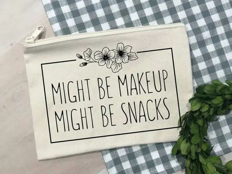 Tan makeup bag with black font that says might be makeup might be snacks with three flowers above. 