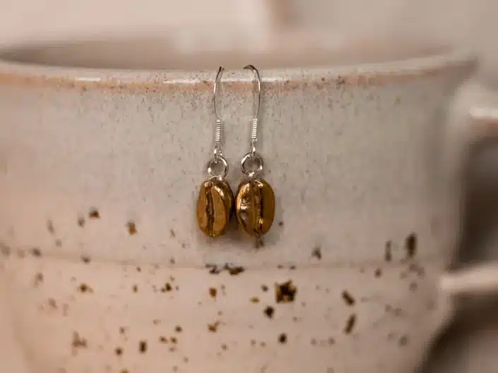 Silver earrings with golden coffee beans on the end hanging off the side of a coffee mug. 