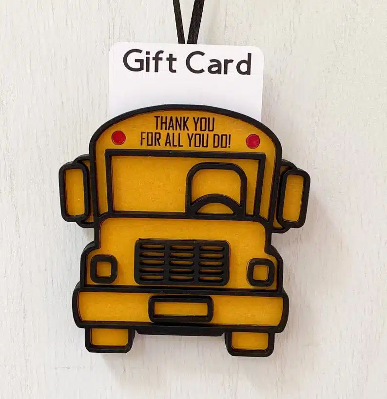 Wooden cut out of a yellow school bus that says thank you for all you do in black print, with a white paper sticking out of the bus ornament that says GIFT CARD. 