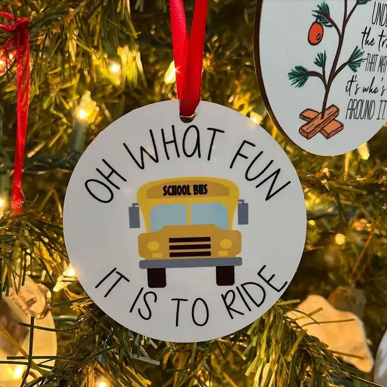 Great Gift Ideas For School Bus Drivers: Close up of a Christmas tree branches with a round white ornament with a yellow school bus on it with black writing around it that says Oh what fun it is to ride. 