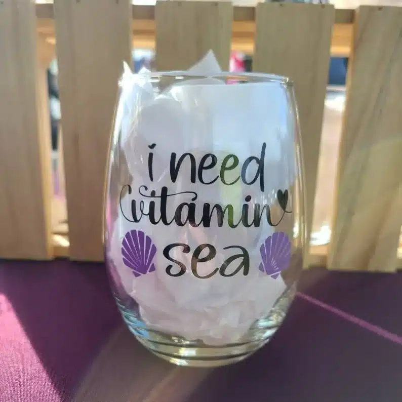 Clear stemless wine glass with black font that says "I need vitamin sea" with two purple shells printed on it. 