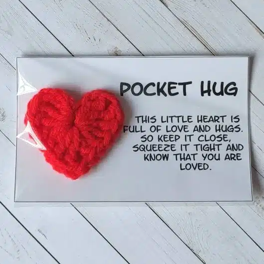 Rectangle white paper with a crocheted red heart with black font that says Pocket hug, this little heart is full of love and hugs. So keep it close squeeze it tight and know that you are loved. 