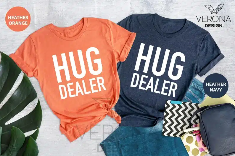 Two t-shirts shown, one coral color and one navy blue both with large white font that says HUG DEALER. 