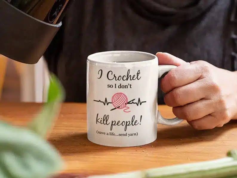 White coffee mug with black font that says "I crochet so I don't kill people!" with a pink yarn on it. 